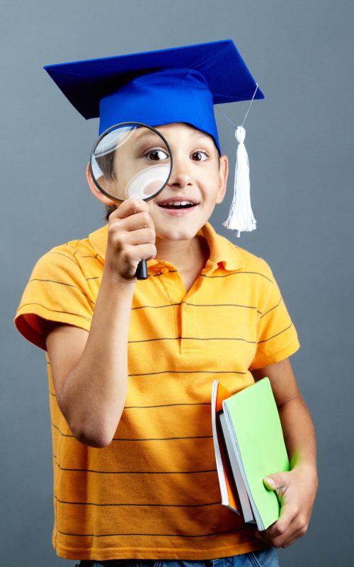Portrait of curious boy in graduation hat looking through magnifying glass